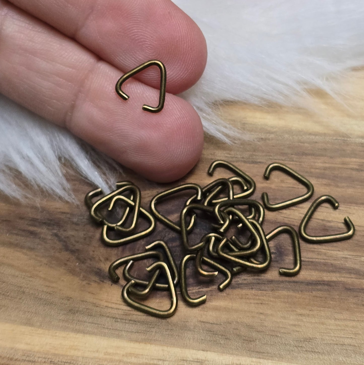 Das Bild zeigt ein Set mit 40 offenen Triangel-Ösen aus antik bronzefarbener Eisenlegierung, die eine Höhe von 9 mm haben. Die Ösen sind in Form eines gleichseitigen Dreiecks gestaltet, wobei eine Seite offen ist, um das Einfügen von Schmuckkomponenten zu erleichtern. Die matte, antik bronzefarbene Oberfläche verleiht den Ösen einen charmanten Vintage-Look, der ideal für verschiedene Schmuckherstellungsprojekte ist. Das Set ist übersichtlich angeordnet und hebt die Vielseitigkeit dieser Ösen hervor, die per