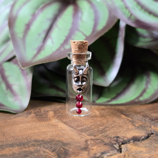 Mini-Glasflasche mit Korken, dekoriert mit Teufelsfratze aus Metall und drei roten Strasssteinen, ideal als Schmuckanhänger oder Sammlerstück.