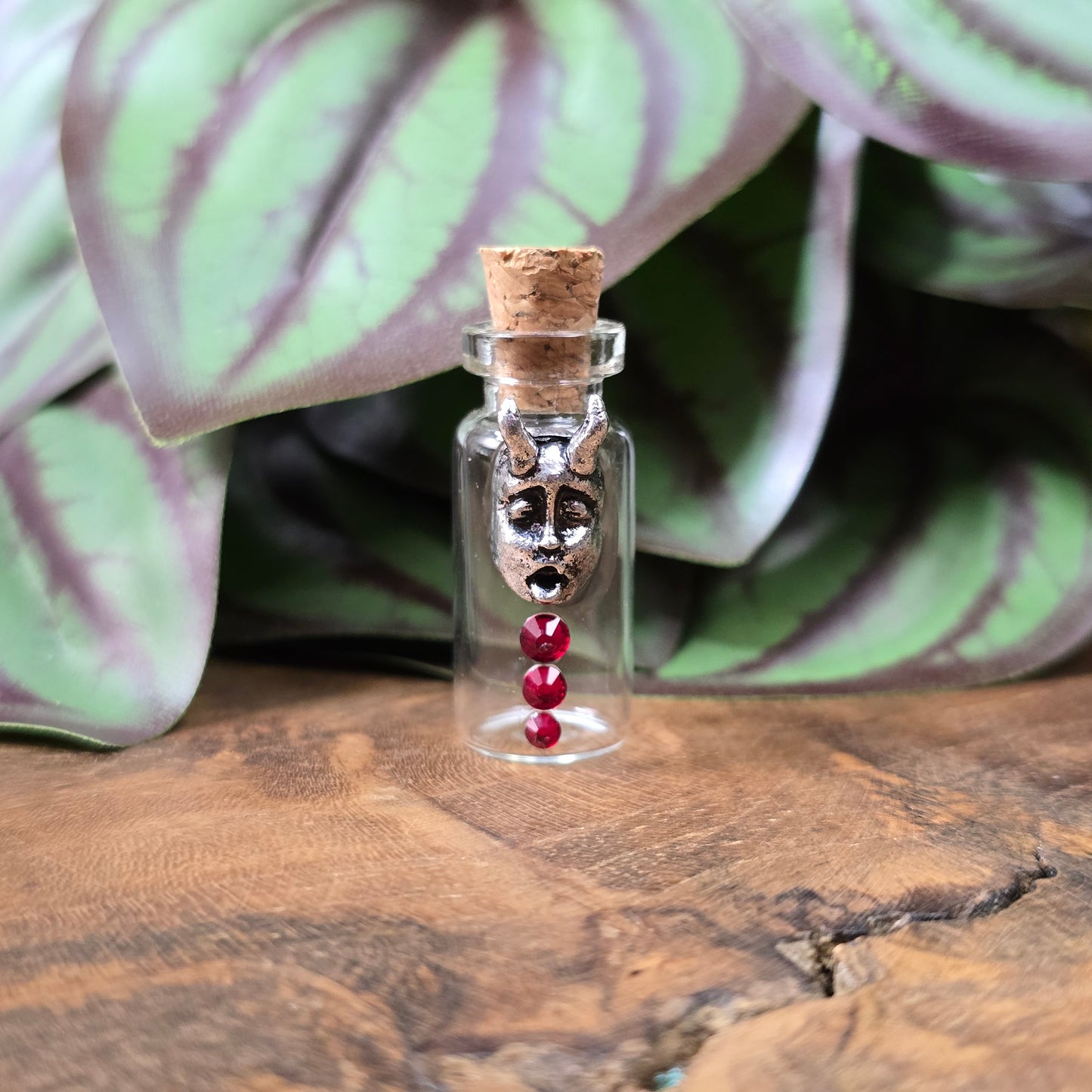 Mini-Glasflasche mit Korken, dekoriert mit Teufelsfratze aus Metall und drei roten Strasssteinen, ideal als Schmuckanhänger oder Sammlerstück.