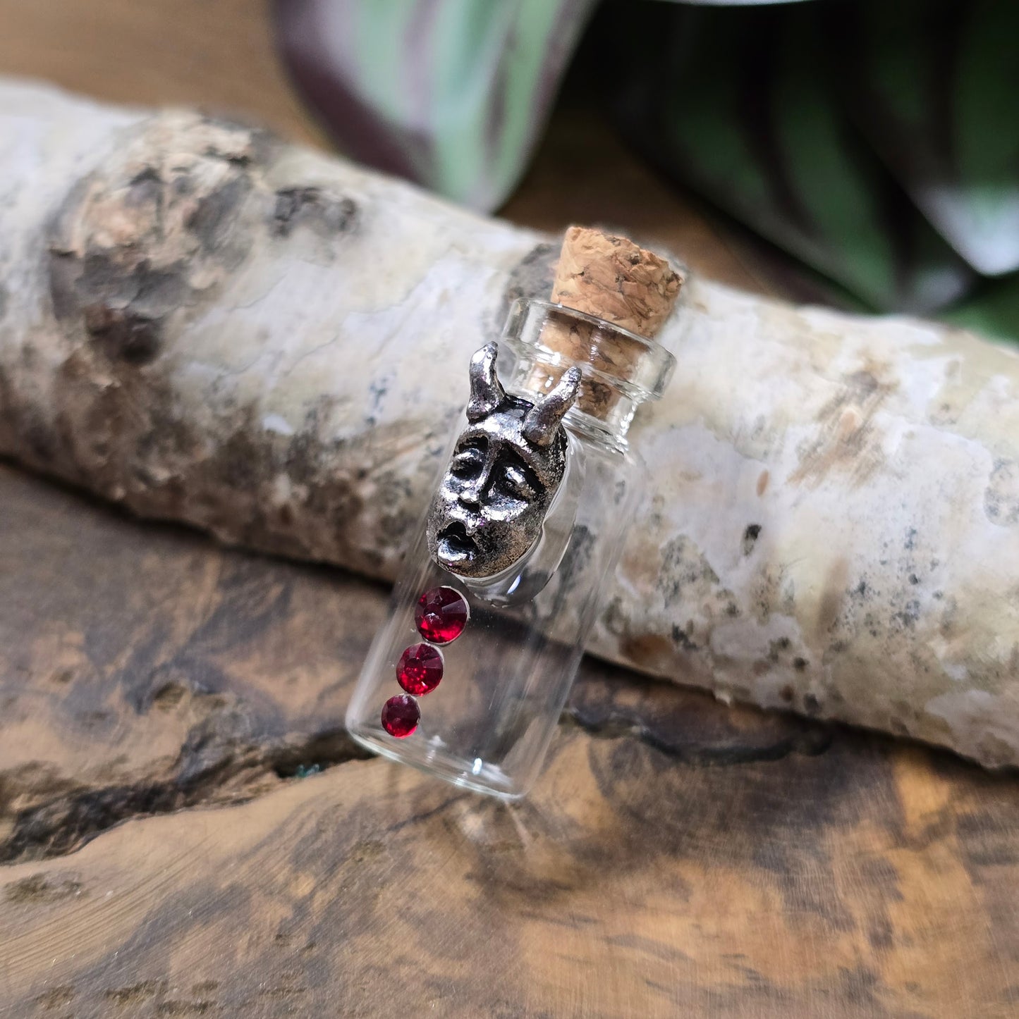 Mini-Glasflasche mit Korken, dekoriert mit Teufelsfratze aus Metall und drei roten Strasssteinen, ideal als Schmuckanhänger oder Sammlerstück.