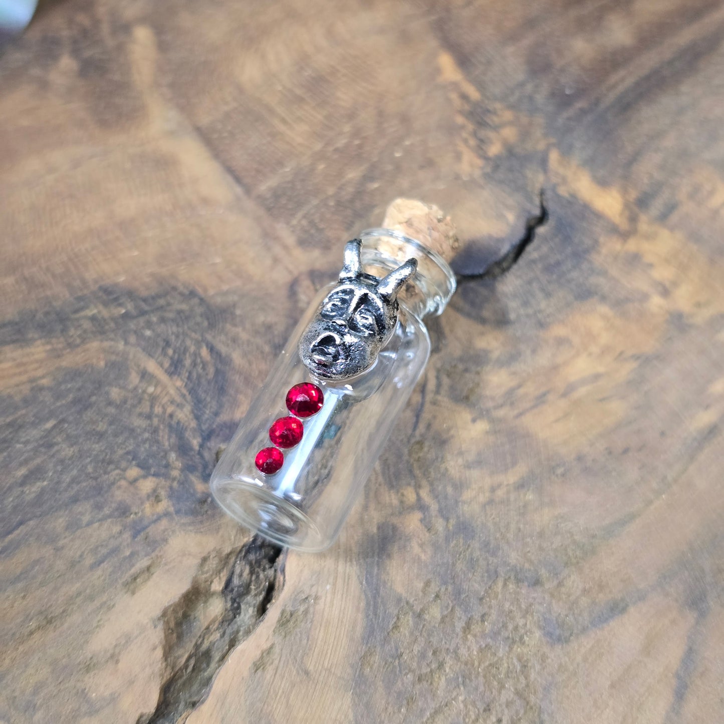Mini-Glasflasche mit Korken, dekoriert mit Teufelsfratze aus Metall und drei roten Strasssteinen, ideal als Schmuckanhänger oder Sammlerstück.