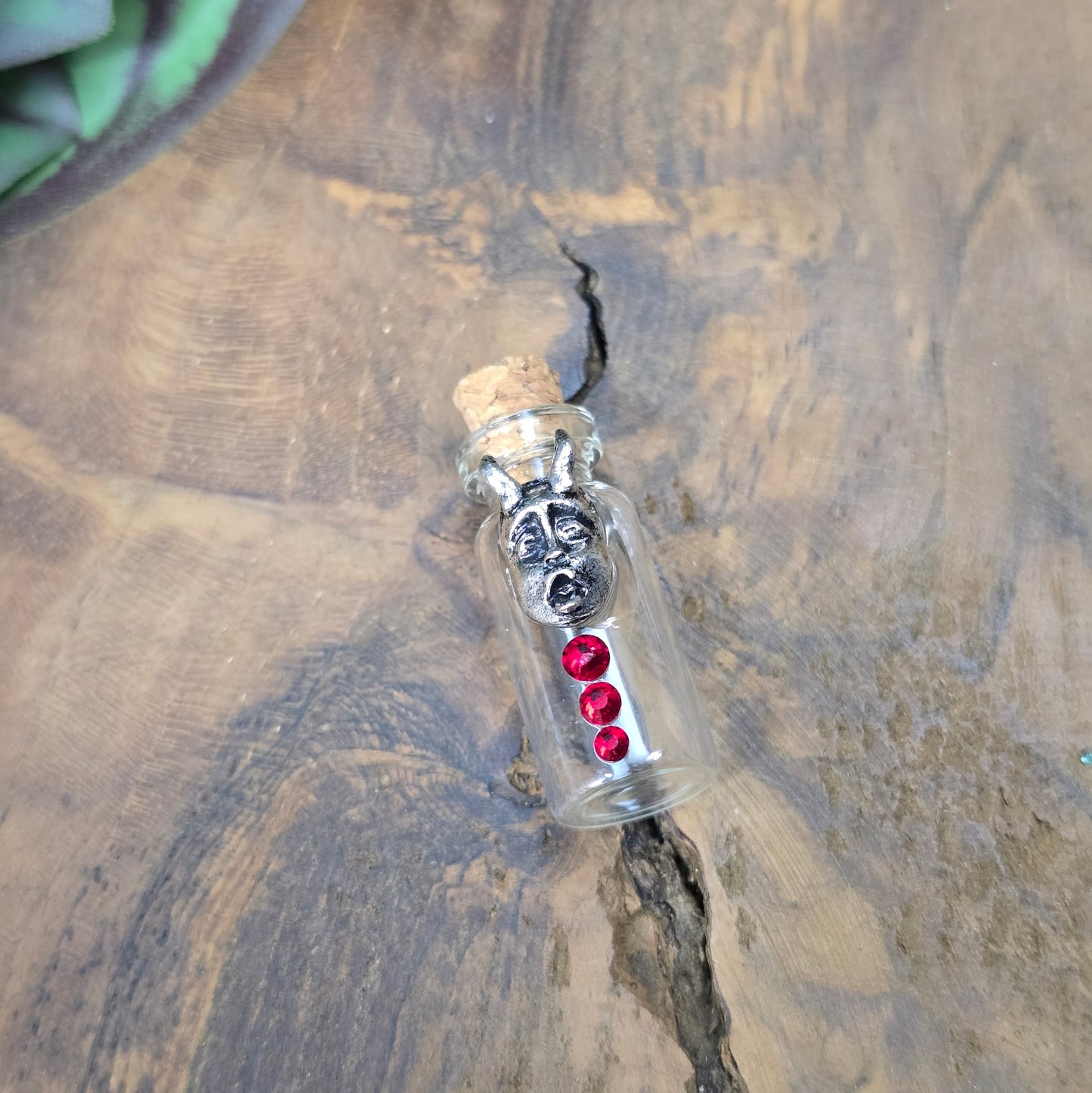 Mini-Glasflasche mit Korken, dekoriert mit Teufelsfratze aus Metall und drei roten Strasssteinen, ideal als Schmuckanhänger oder Sammlerstück.