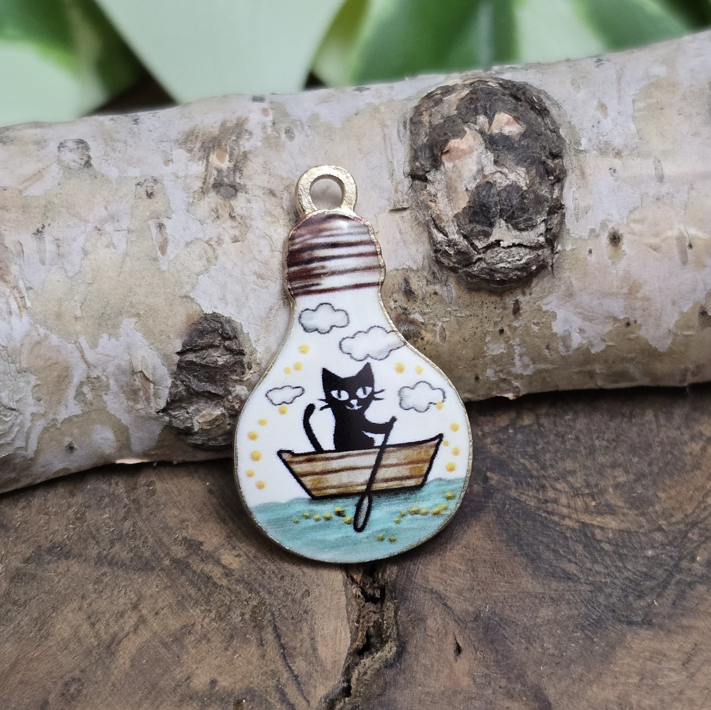 Das Bild zeigt einen goldfarbenen Anhänger in Glühbirnenform, auf dem ein Katzenmotiv abgebildet ist. Eine Katze sitzt in einem Ruderboot und fährt auf einem malerischen See. Das filigrane Design ist von einer goldenen Umrandung eingefasst, die dem Anhänger eine edle Note verleiht. Der Anhänger hat eine Größe von 27mm und eignet sich hervorragend für Schmuckherstellung oder DIY-Projekte.