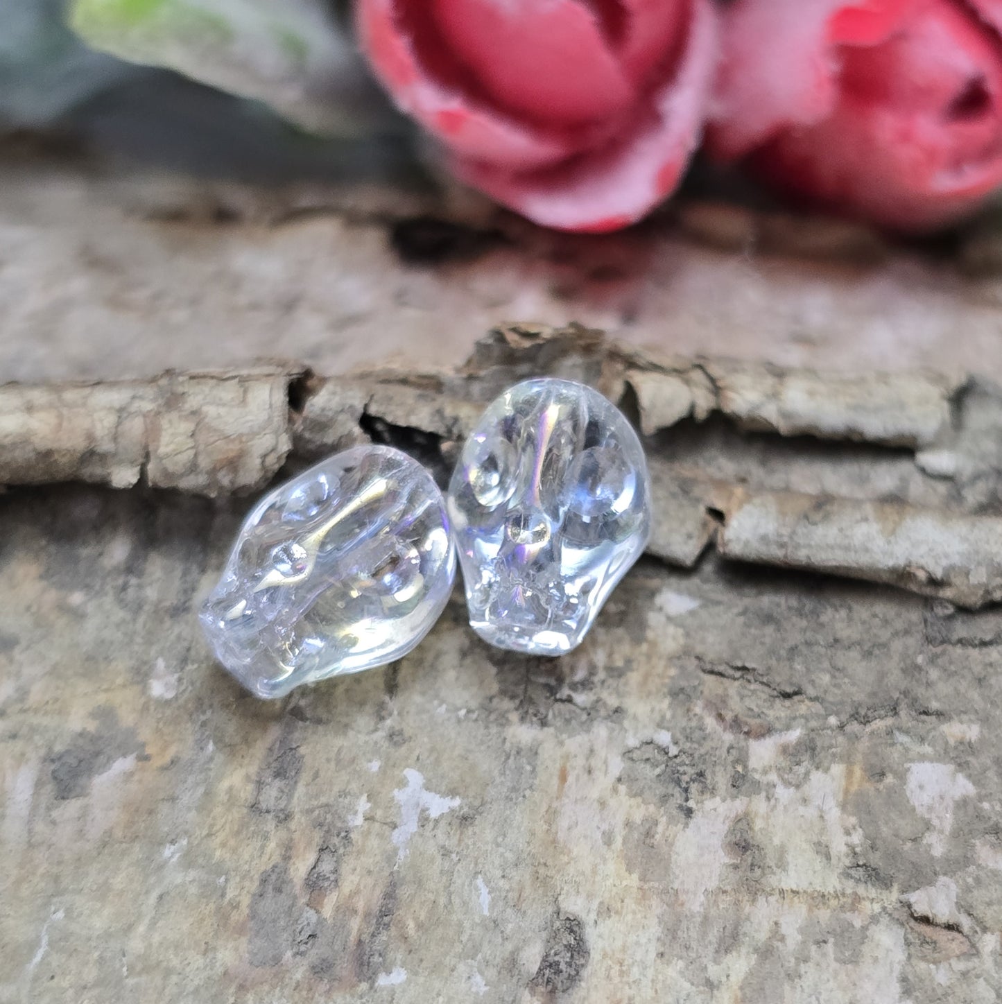 Das Bild zeigt zwei schimmernde Glasperlen in Totenkopf-Form mit Crystal-AB-Beschichtung. Die Perlen sind dreidimensional gestaltet, sodass Details wie Augenhöhlen, Nase und Kiefer deutlich erkennbar sind. Ihre Oberfläche reflektiert das Licht in irisierenden Farben, die je nach Blickwinkel changieren. Die Bohrung verläuft von oben nach unten, sodass sie ideal für Schmuckprojekte geeignet sind.