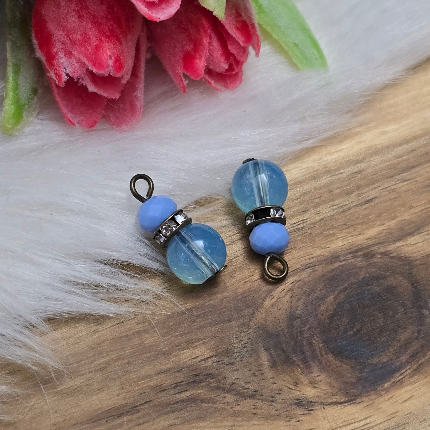 Das Bild zeigt zwei handgefertigte Anhänger mit einer Kombination aus transparenten hellblauen Glasperlen und opaken hellblauen Kristallglasperlen. Ein funkelndes Strassrondell setzt glitzernde Akzente, während antik bronzefarbige Metallelemente dem Design einen charmanten Vintage-Touch verleihen. Die filigranen Details und die harmonische Farbzusammenstellung machen die Anhänger zu einem stilvollen Blickfang für Schmuckkreationen.