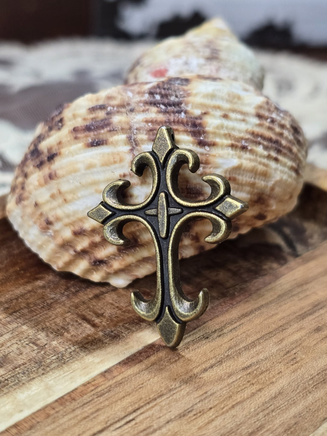 Barocker Kreuz-Anhänger 32x24mm in Antikbronze dekoriert vor einer Muschel auf Holz.