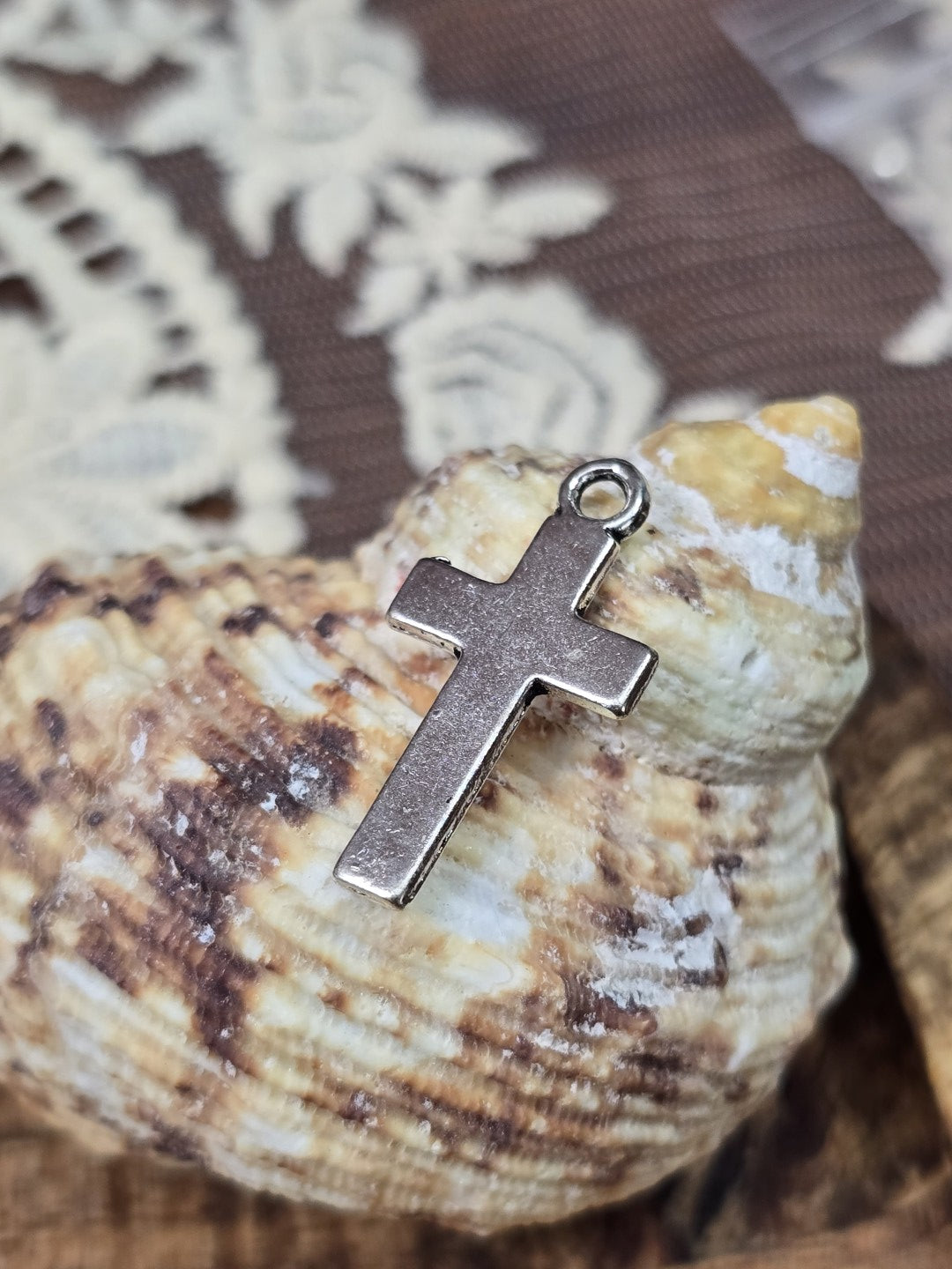 Silberner Kreuz-Schmuckanhänger mit Öse aufgestellt vor einer Muschel.