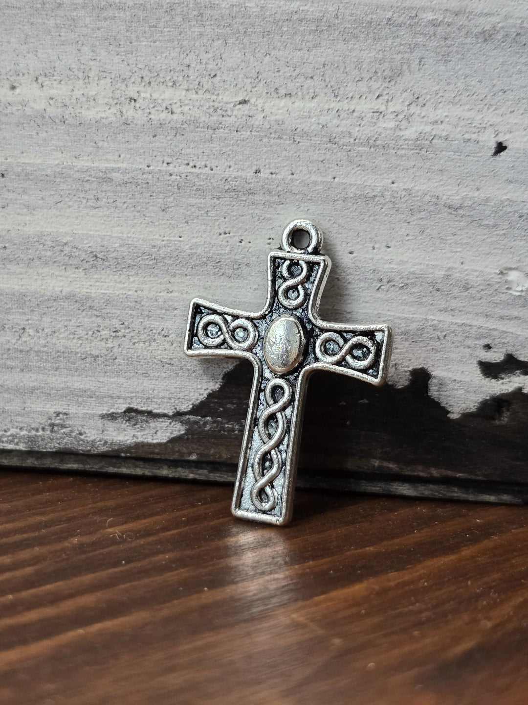 Kreuz-Anhänger in Antiksilber mit Flechtmuster aufrecht stehend vor rustikalem Holz-Hintergrund.