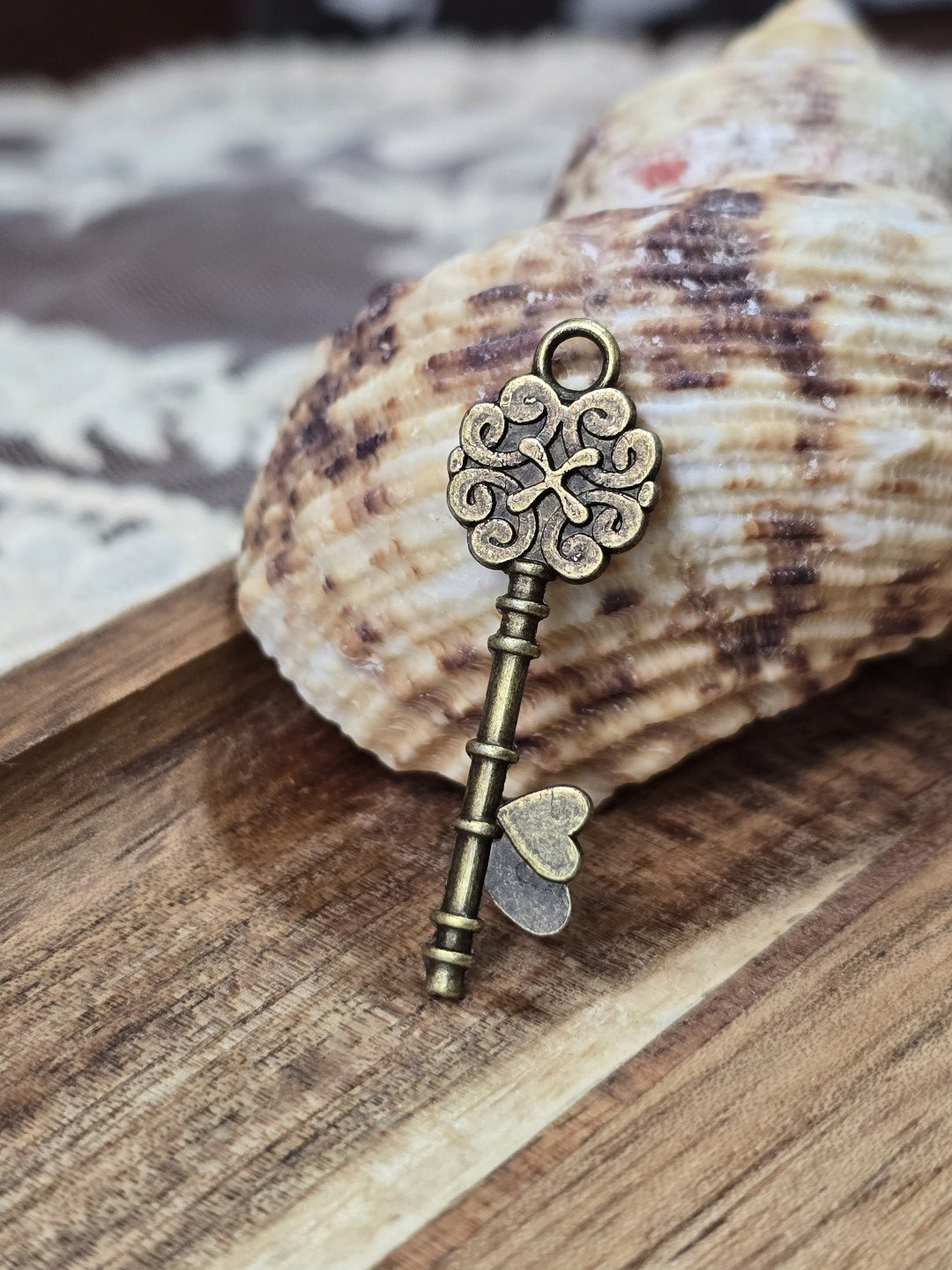 Schlüssel-Schmuckanhänger in Bronze-Optik mit Öse dekoriert vor einer Muschel auf Holz.