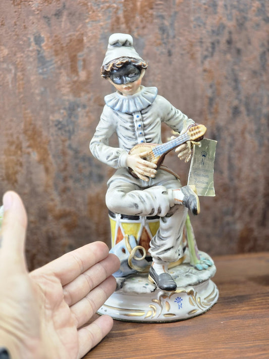 Vintage Porzellanfigur mit Harlekin, Mandoline und schwarzer Augenmaske, neben einer Hand zur Größenwirkung auf Holztisch fotografiert.