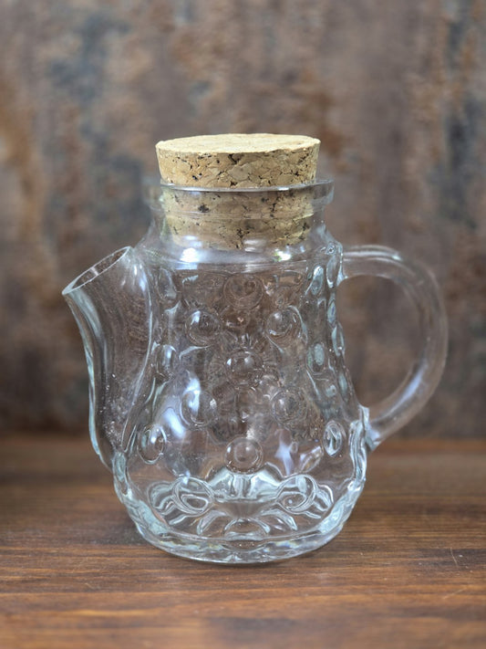 Vintage Erdnussspender aus transparentem Pressglas mit Korken, geschwungenem Henkel und kurzem Ausguss in gerader Vorderansicht