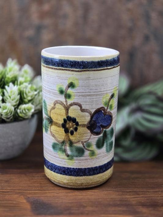 Vintage Keramikvase mit floralem Dekor in Gelb, Blau und Grün, zylindrischer Form und heller Innenwand vor Pflanzenkulisse.
