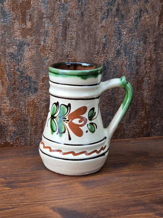 Vintage Keramikkrug mit floralem Dekor in Grün und Braun, cremefarbener Glasur, grünem Henkel und sichtbarem rotbrennendem Keramikscherben am Boden.