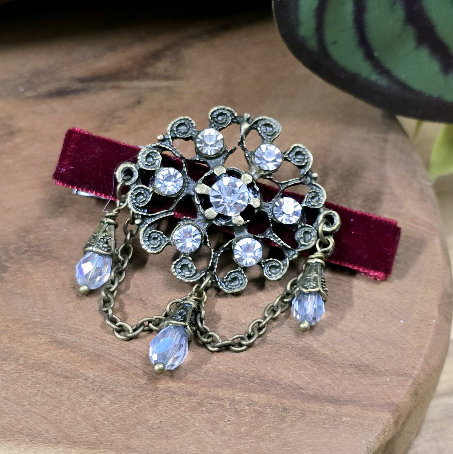Das Bild zeigt eine Krokodilspange im Vintagelook mit Strass und dunkelrotem Samt, Kristallglasanhänger, Kettchen, Einzelstück
Lass dich von der zeitlosen Eleganz vergangener Epochen verzaubern mit dieser exquisiten Krokodilspange im Vintagestil. Sie ist kunstvoll verziert mit einem filigranen, antik bronzefarbigen Schmuckteil, welches mit funkelndem Strass besetzt ist.