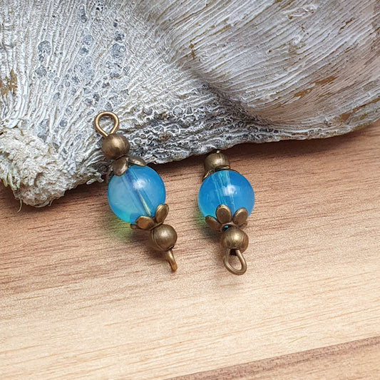 Das Bild zeigt zwei handgefertigte Verbinder mit transparenten, aquablauen Glasperlen, die in antik bronzefarbige Metallelemente eingefasst sind. Die Perlen schimmern sanft und verleihen den Verbindern eine edle Optik. Die filigrane Metallverzierung betont den Vintage-Charakter der Schmuckelemente. Mit einer Länge von ca. 24mm sind sie vielseitig einsetzbar – perfekt für Armbänder, Ketten oder Ohrringe.
