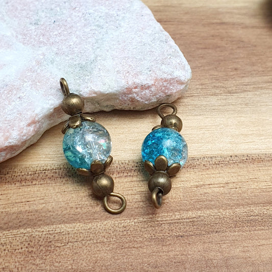 Zwei handgefertigte Schmuckverbinder mit aquablauen Crash-Glasperlen und antik bronzefarbenen Metallelementen liegen nebeneinander auf einer neutralen Oberfläche. Die Glasperlen haben eine gesprungene Optik, die das Licht reflektiert und für einen besonderen Schimmereffekt sorgt. Die bronzefarbene Fassung verleiht den Verbindern einen edlen Vintage-Look. Durch die Ösen an beiden Enden eignen sie sich ideal für Schmuckprojekte wie Armbänder, Halsketten oder Ohrringe. ✨