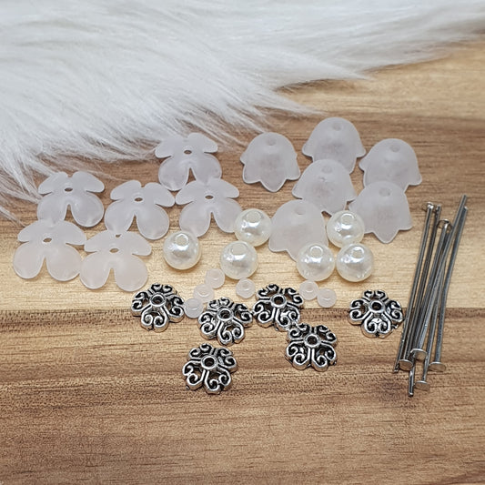 Das Bild zeigt ein umfangreiches Bastelset zur Herstellung von 6 kleinen Anhängern. Im Set sind verschiedene Materialien angeordnet, darunter orange matte Glasperlen, cremeweiße Acrylperlen, Blütenkäppchen in der gleichen Farbe und antik silberfarbige Sternkäppchen. Außerdem sind Prismenstifte zu sehen, die für die Verbindung der einzelnen Komponenten verwendet werden. Alle Teile sind sorgfältig angeordnet, um die kreativen Möglichkeiten zu verdeutlichen, die das Set bietet. Die Darstellung vermittelt einen