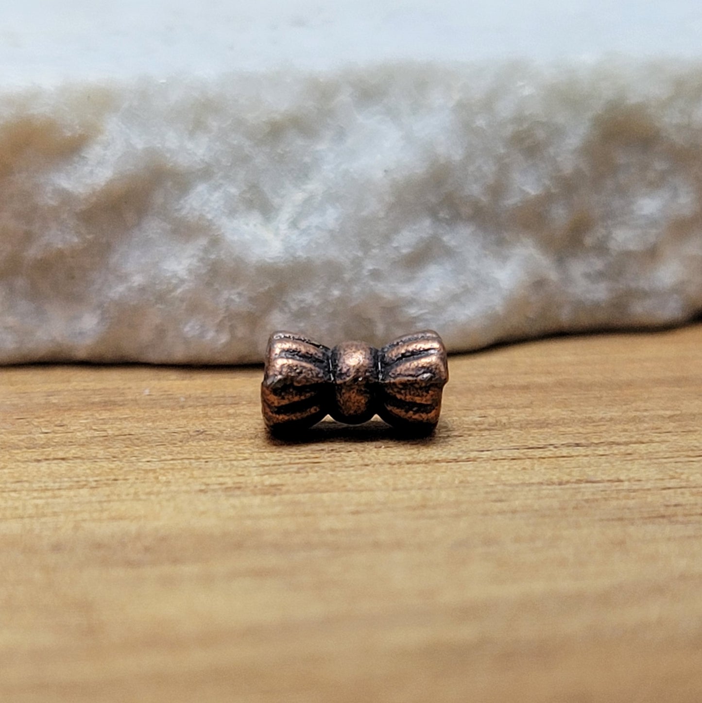 Diese Packung enthält 10 kleine Metallperlen in antik kupferfarbig.  Die Perlen haben die Form eines kleinen aufgeplusterten Schleifchens. Das Fädelloch der Perlen verläuft durch den langen Teil der Perle.