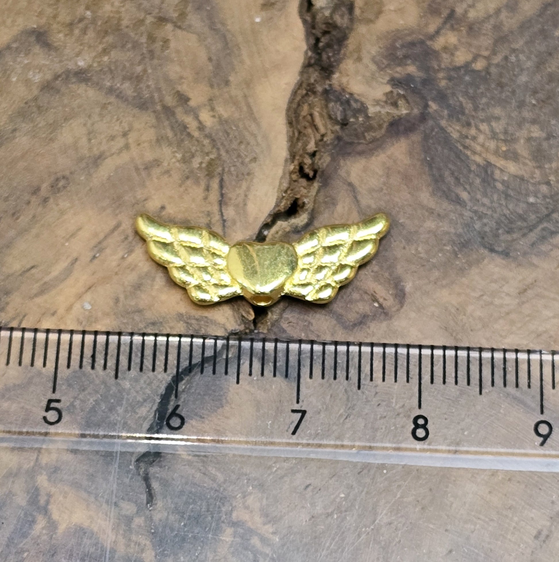 Das Bild zeigt vergoldete Metallperlen in Form von Engelsflügeln mit einem Herz in der Mitte. Die Perlen sind 22 mm groß und eignen sich für die Herstellung von Schutzengel-Anhängern oder für DIY-Schmuckprojekte. Sie haben eine glänzende, goldene Oberfläche und ein elegantes Design.
