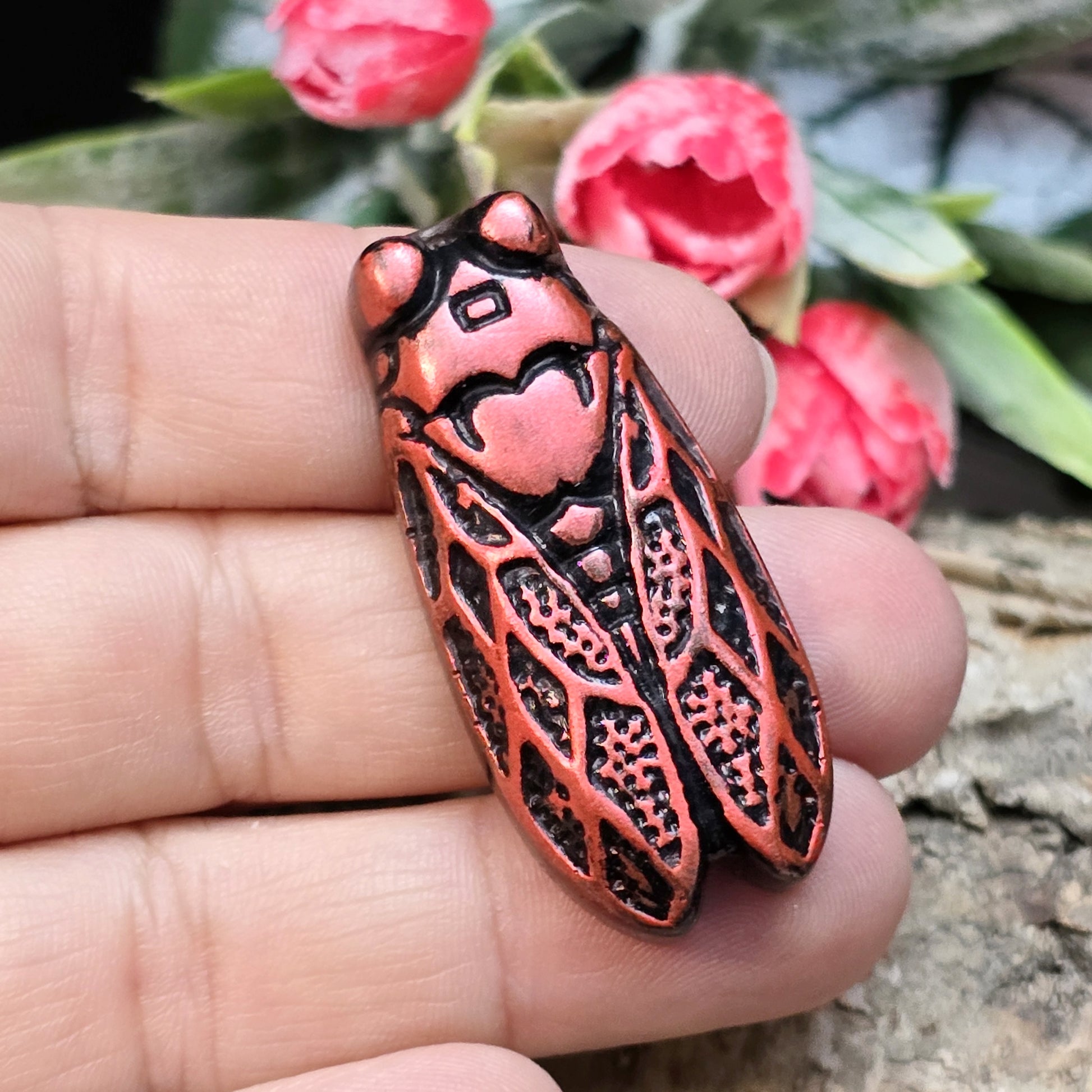 Das Bild zeigt einen handgefertigten Zikaden-Cabochon aus Kunstharz in einer eleganten Karminrot-Optik. Die Zikade hat eine flache Rückseite mit einem kleinen Loch zwischen den Augen, das sich perfekt für die Anbringung einer Anhängeröse eignet.