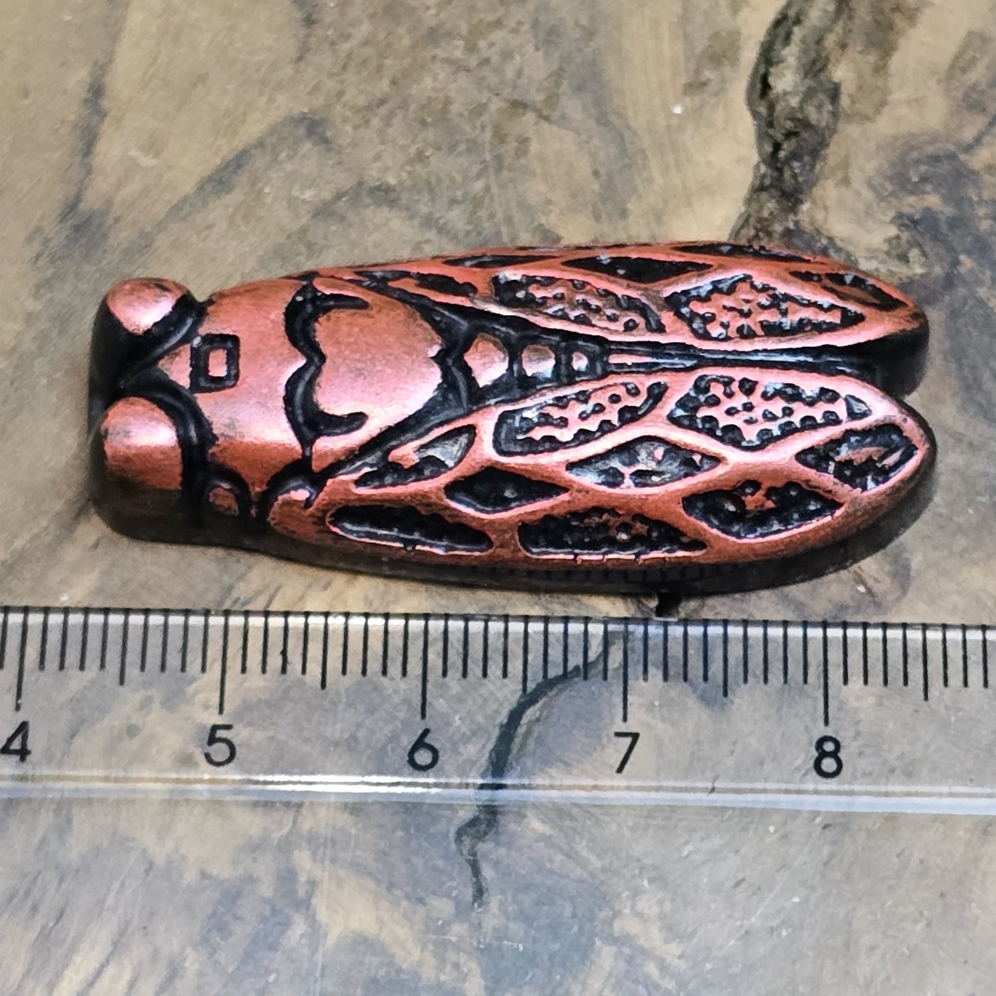 Das Bild zeigt einen handgefertigten Zikaden-Cabochon aus Kunstharz in einer eleganten Karminrot-Optik. Die Zikade hat eine flache Rückseite mit einem kleinen Loch zwischen den Augen, das sich perfekt für die Anbringung einer Anhängeröse eignet.