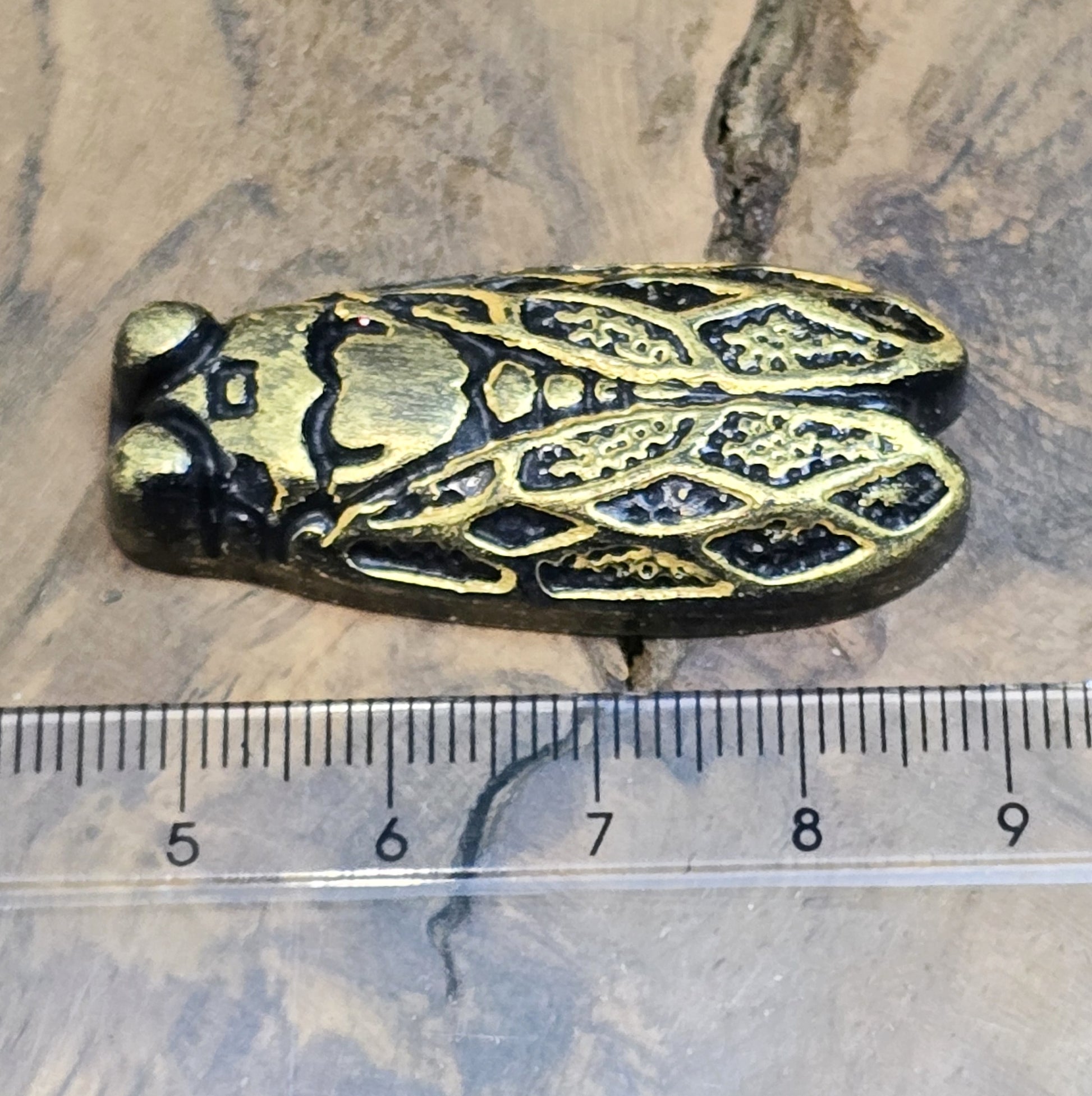 Das Bild zeigt einen handgefertigten Zikaden-Cabochon aus Kunstharz in einer edlen Bronze-Optik. Die Zikade hat eine flache Rückseite mit einem kleinen Loch zwischen den Augen, ideal für die Anbringung einer Anhängeröse.