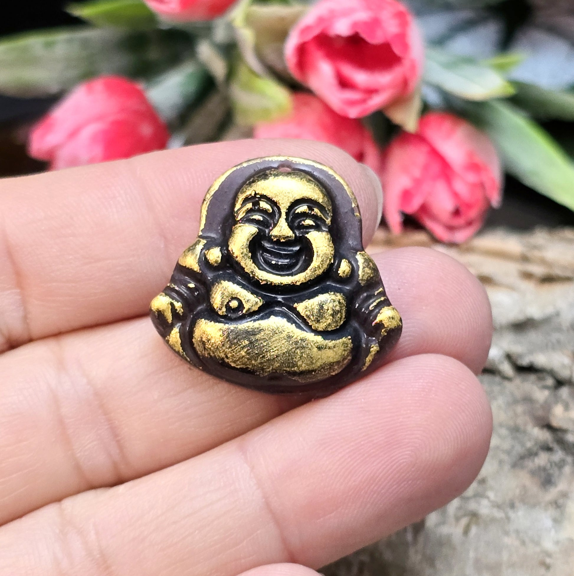 Das Bild zeigt einen handgefertigten Cabochon in Form eines lachenden Buddhas aus Kunstharz. Der Buddha hat eine schwarze Oberfläche mit bronzefarbenen Details und ein kleines Loch am oberen Rand. Die Rückseite ist flach, ideal für die Verwendung als Anhänger oder in Bastelprojekten.