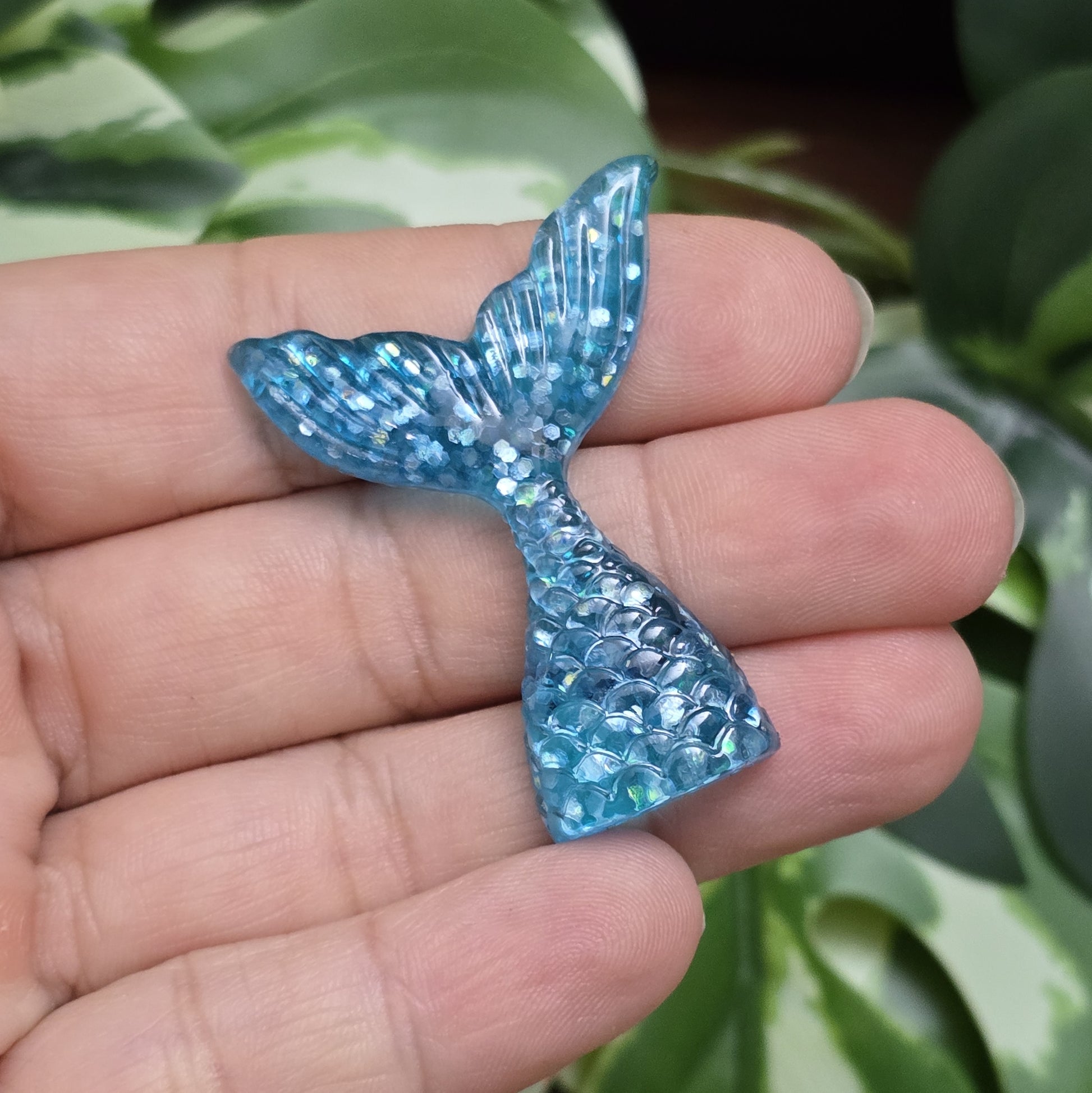 Das Bild zeigt einen handgefertigten Cabochon in Form einer Meerjungfrauenschwanzflosse. Der Anhänger besteht aus Kunstharz und leuchtet in einem schimmernden Aqua Blau mit feinem Glitter. Die geschwungene Form und die detailreiche Struktur verleihen ihm eine elegante, maritime Optik.