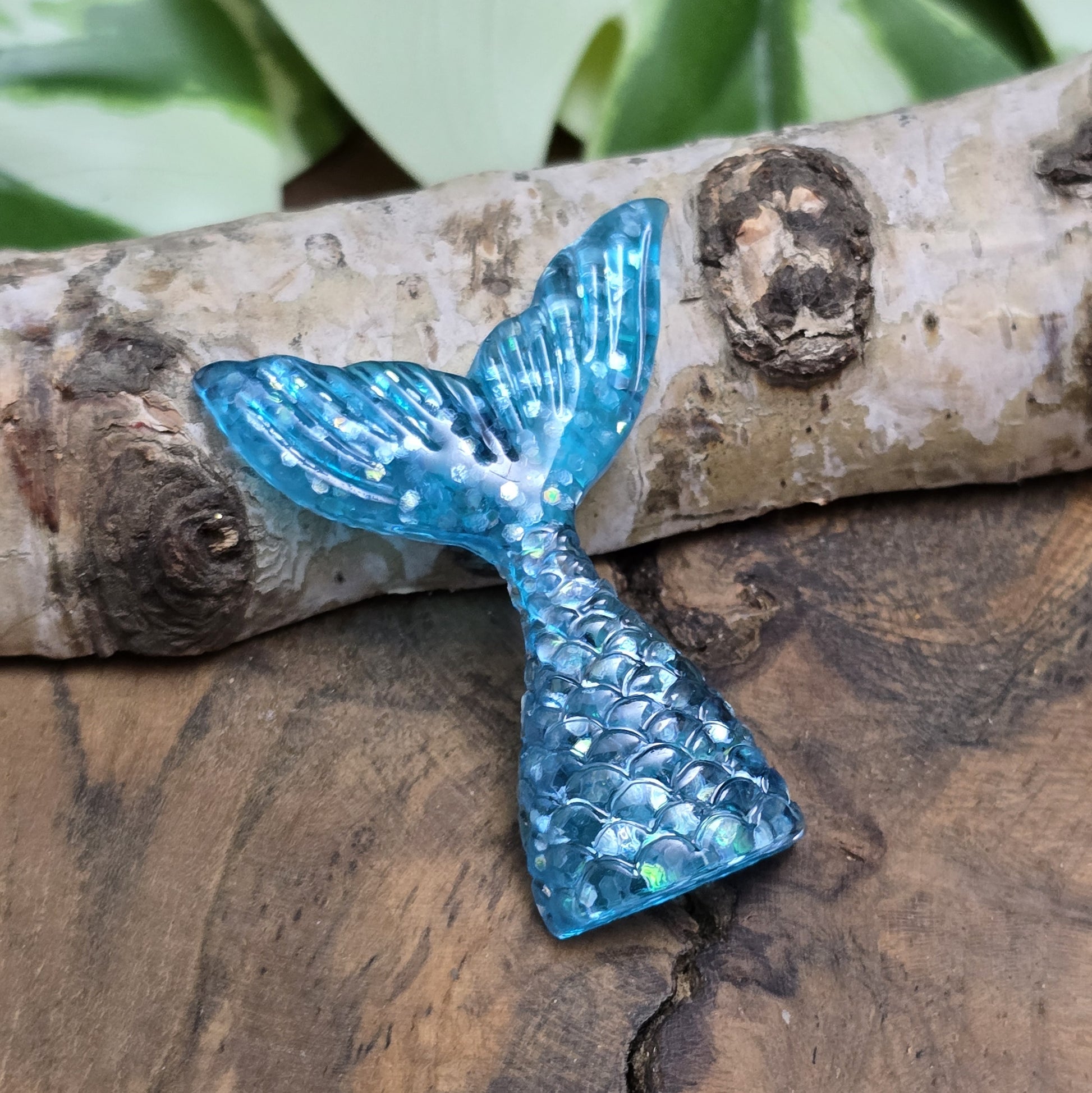 Das Bild zeigt einen handgefertigten Cabochon in Form einer Meerjungfrauenschwanzflosse. Der Anhänger besteht aus Kunstharz und leuchtet in einem schimmernden Aqua Blau mit feinem Glitter. Die geschwungene Form und die detailreiche Struktur verleihen ihm eine elegante, maritime Optik.
