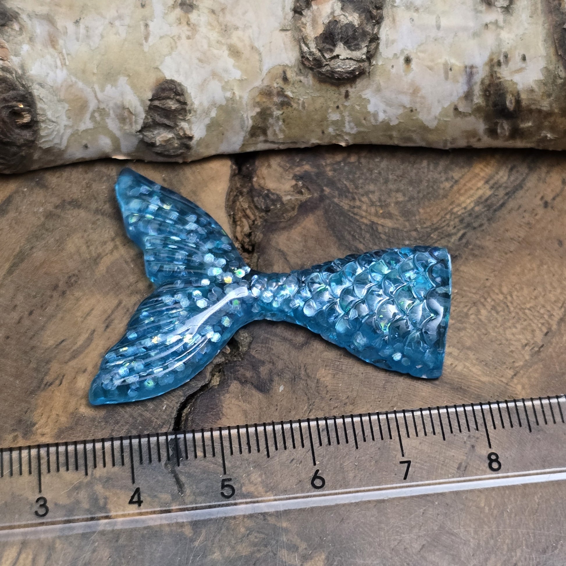 Das Bild zeigt einen handgefertigten Cabochon in Form einer Meerjungfrauenschwanzflosse. Der Anhänger besteht aus Kunstharz und leuchtet in einem schimmernden Aqua Blau mit feinem Glitter. Die geschwungene Form und die detailreiche Struktur verleihen ihm eine elegante, maritime Optik.