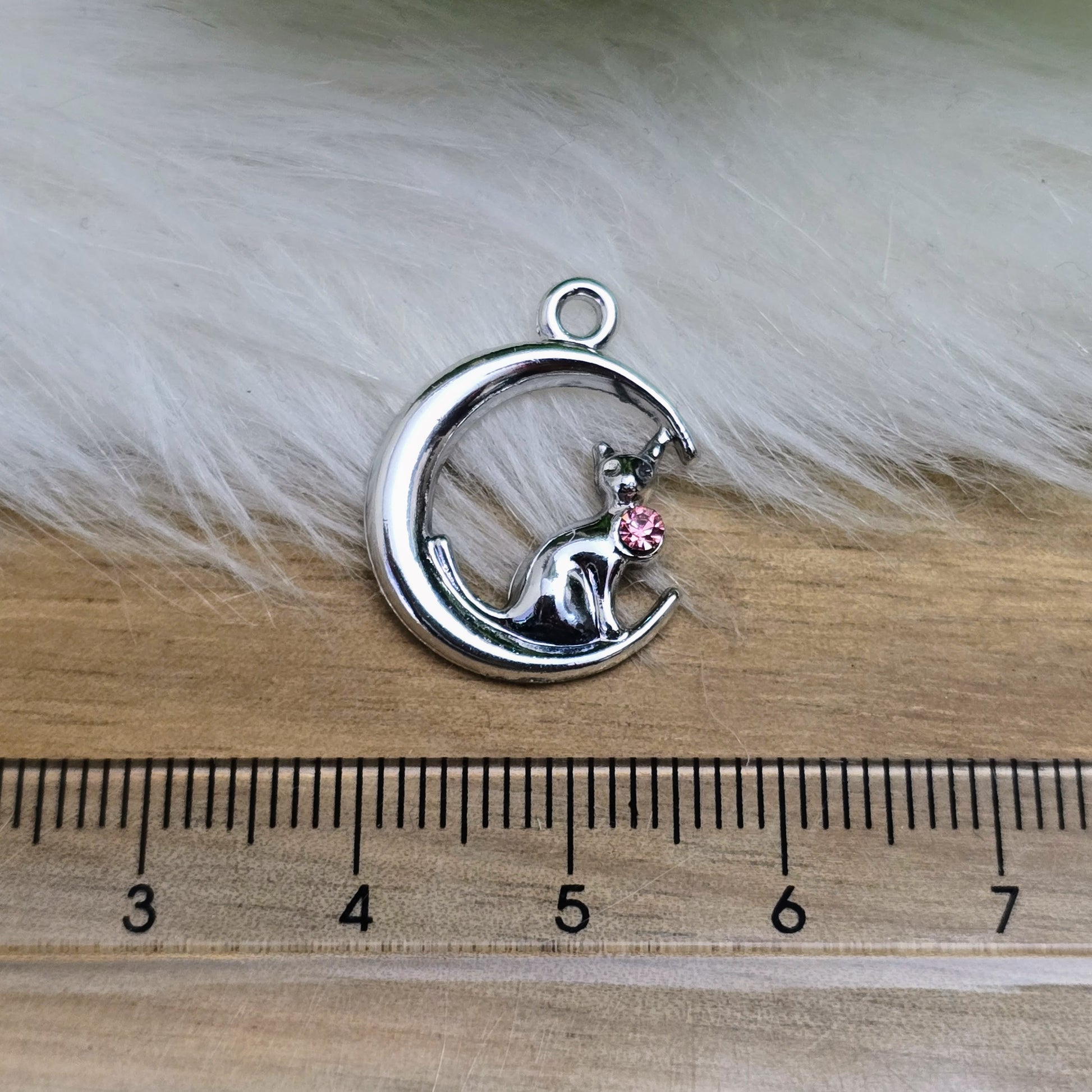 Das Bild zeigt einen Anhänger in Silberfarbe, der eine Katze im Mond darstellt. Die Katze ist elegant im Mond sitzend, begleitet von einem funkelnden rosé Strass-Stein. Der Anhänger ist ca. 20mm groß und eignet sich perfekt für die Schmuckherstellung oder als kreatives Accessoire.
