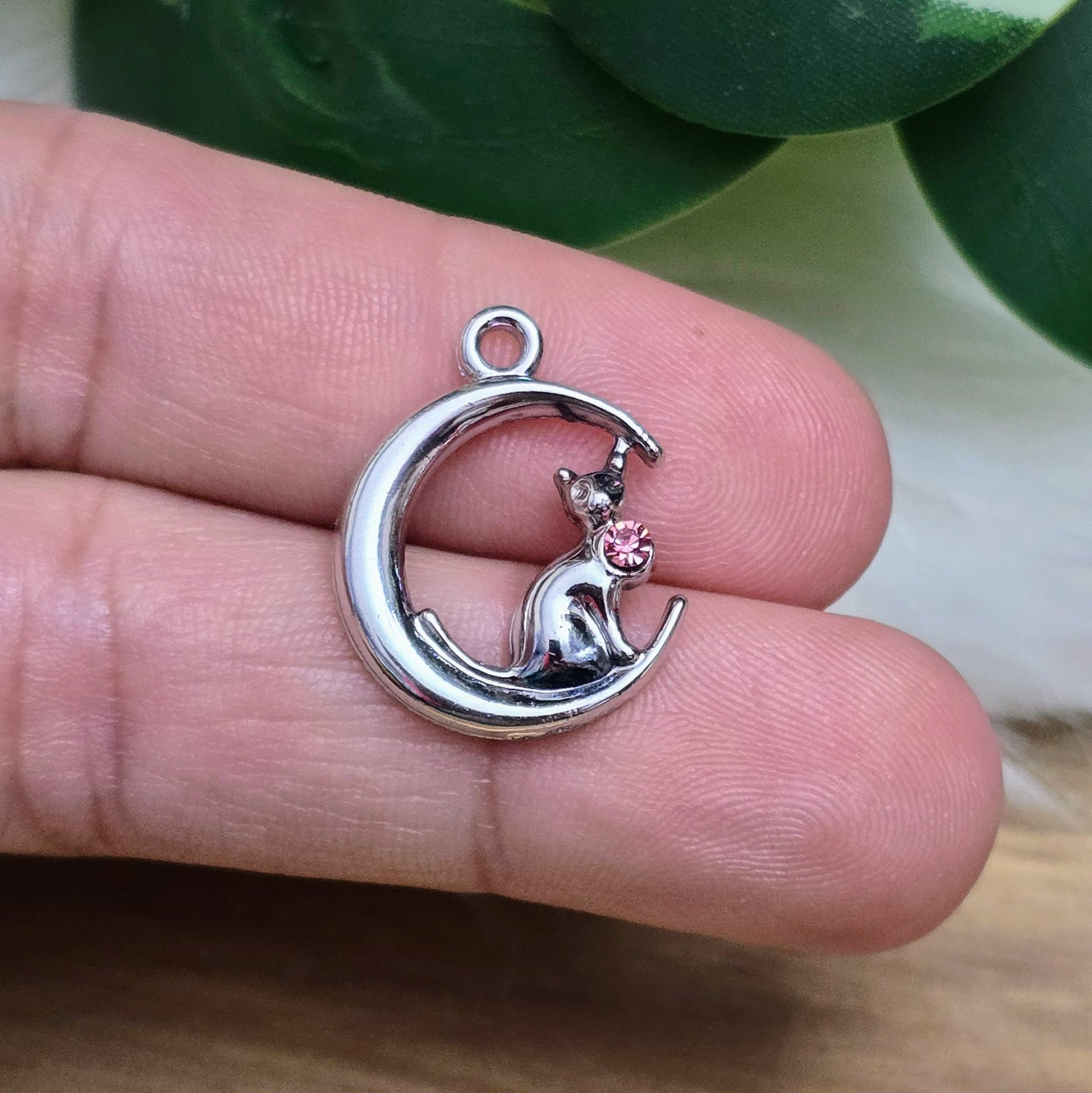 Das Bild zeigt einen Anhänger in Silberfarbe, der eine Katze im Mond darstellt. Die Katze ist elegant im Mond sitzend, begleitet von einem funkelnden rosé Strass-Stein. Der Anhänger ist ca. 20mm groß und eignet sich perfekt für die Schmuckherstellung oder als kreatives Accessoire.