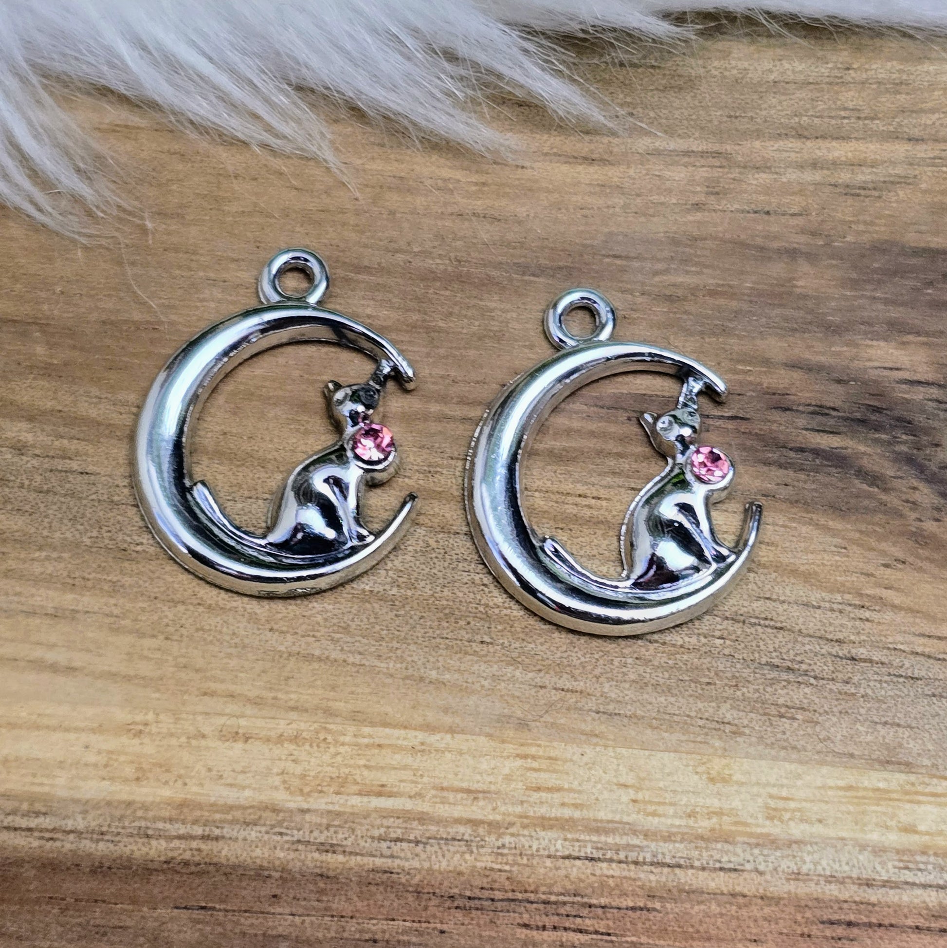 Das Bild zeigt einen Anhänger in Silberfarbe, der eine Katze im Mond darstellt. Die Katze ist elegant im Mond sitzend, begleitet von einem funkelnden rosé Strass-Stein. Der Anhänger ist ca. 20mm groß und eignet sich perfekt für die Schmuckherstellung oder als kreatives Accessoire.