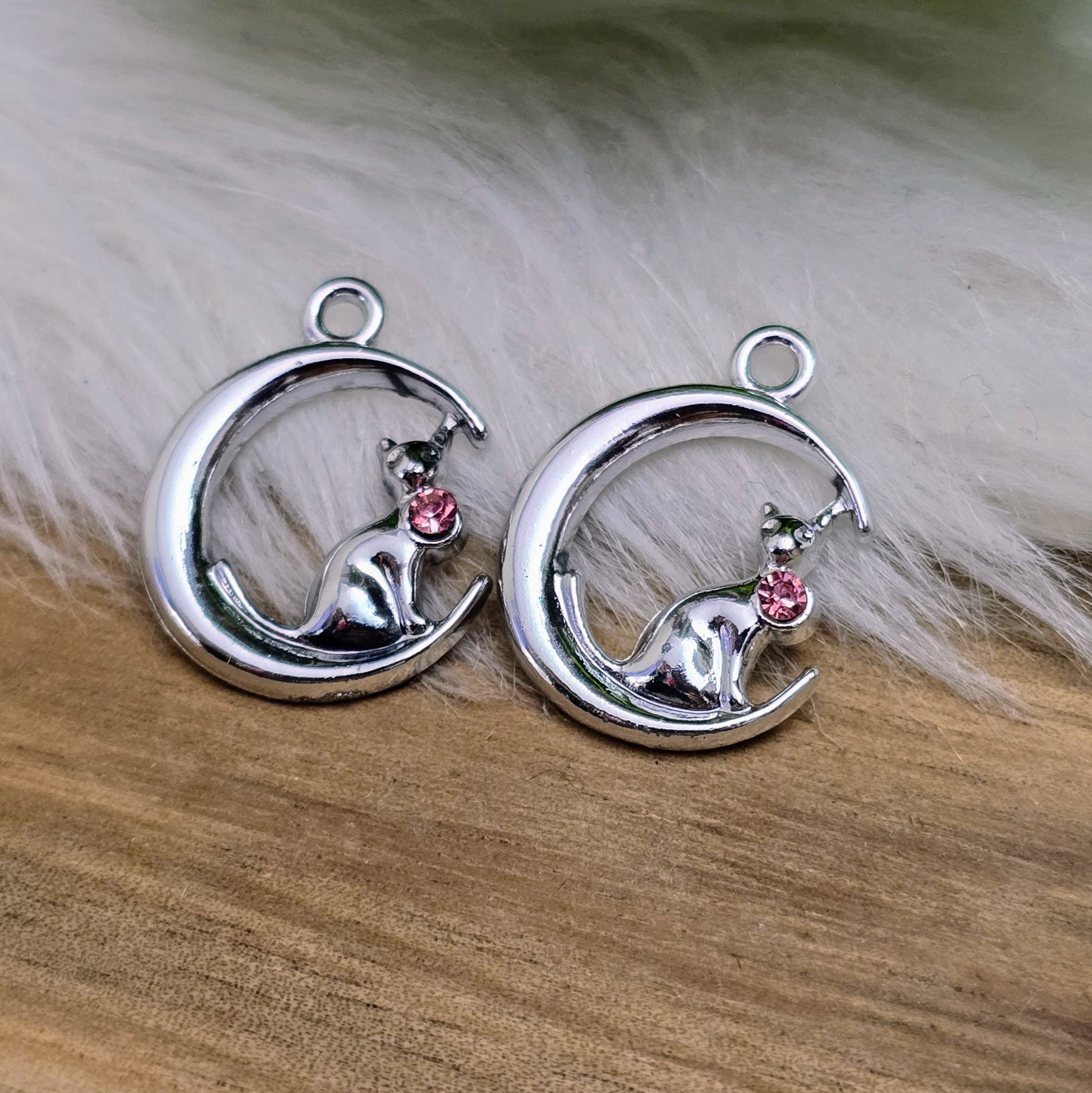 Das Bild zeigt einen Anhänger in Silberfarbe, der eine Katze im Mond darstellt. Die Katze ist elegant im Mond sitzend, begleitet von einem funkelnden rosé Strass-Stein. Der Anhänger ist ca. 20mm groß und eignet sich perfekt für die Schmuckherstellung oder als kreatives Accessoire.