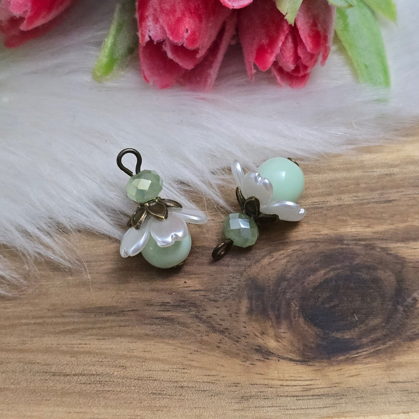 Das Bild zeigt zwei handgefertigte Anhänger mit lindgrünen Glasperlen. Jede Perle ist von einem perlweißen Blütenkäppchen aus Acryl umgeben, während eine facettierte Kristallglasperle in Lindgrün für funkelnde Akzente sorgt. Eine antik bronzefarbene Perlenkappe verleiht dem Design einen nostalgischen Charme. Die Anhänger haben eine Größe von ca. 20mm und eignen sich ideal für Schmuckkreationen im Vintage- oder romantischen Stil.