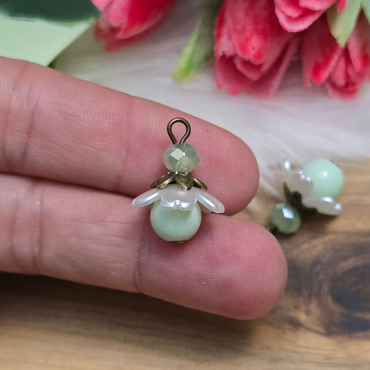 Das Bild zeigt zwei handgefertigte Anhänger mit lindgrünen Glasperlen. Jede Perle ist von einem perlweißen Blütenkäppchen aus Acryl umgeben, während eine facettierte Kristallglasperle in Lindgrün für funkelnde Akzente sorgt. Eine antik bronzefarbene Perlenkappe verleiht dem Design einen nostalgischen Charme. Die Anhänger haben eine Größe von ca. 20mm und eignen sich ideal für Schmuckkreationen im Vintage- oder romantischen Stil.
