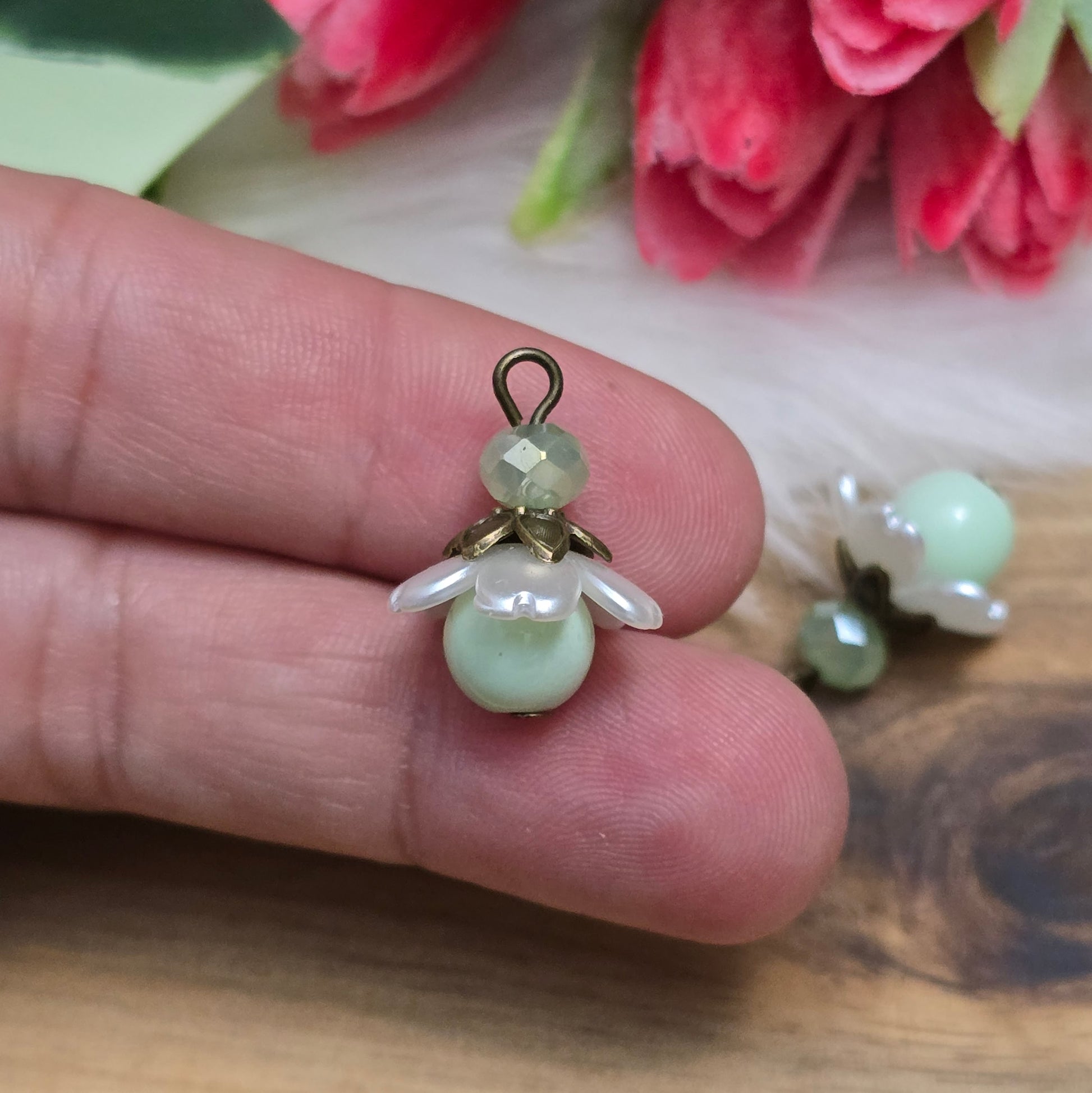 Das Bild zeigt zwei handgefertigte Anhänger mit lindgrünen Glasperlen. Jede Perle ist von einem perlweißen Blütenkäppchen aus Acryl umgeben, während eine facettierte Kristallglasperle in Lindgrün für funkelnde Akzente sorgt. Eine antik bronzefarbene Perlenkappe verleiht dem Design einen nostalgischen Charme. Die Anhänger haben eine Größe von ca. 20mm und eignen sich ideal für Schmuckkreationen im Vintage- oder romantischen Stil.