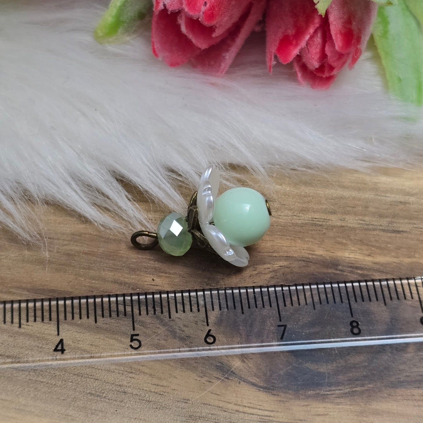Das Bild zeigt zwei handgefertigte Anhänger mit lindgrünen Glasperlen. Jede Perle ist von einem perlweißen Blütenkäppchen aus Acryl umgeben, während eine facettierte Kristallglasperle in Lindgrün für funkelnde Akzente sorgt. Eine antik bronzefarbene Perlenkappe verleiht dem Design einen nostalgischen Charme. Die Anhänger haben eine Größe von ca. 20mm und eignen sich ideal für Schmuckkreationen im Vintage- oder romantischen Stil.