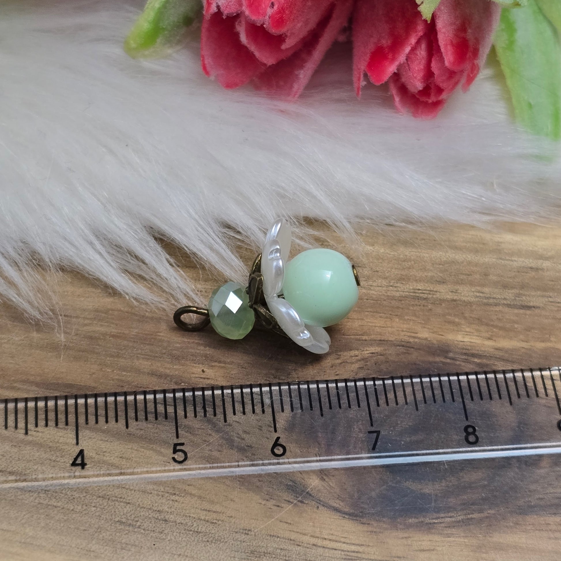 Das Bild zeigt zwei handgefertigte Anhänger mit lindgrünen Glasperlen. Jede Perle ist von einem perlweißen Blütenkäppchen aus Acryl umgeben, während eine facettierte Kristallglasperle in Lindgrün für funkelnde Akzente sorgt. Eine antik bronzefarbene Perlenkappe verleiht dem Design einen nostalgischen Charme. Die Anhänger haben eine Größe von ca. 20mm und eignen sich ideal für Schmuckkreationen im Vintage- oder romantischen Stil.