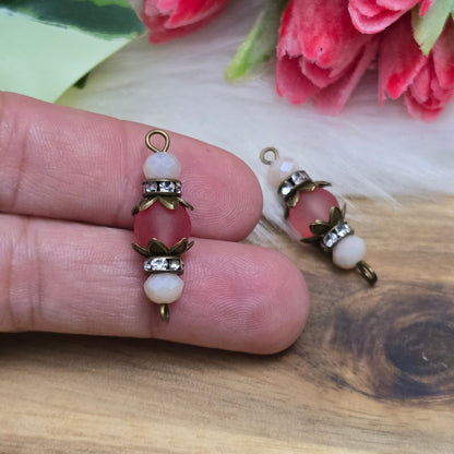 Das Bild zeigt zwei handgefertigte Verbinder im Shabby-Stil, die aus matten, transparenten Glasperlen in Lachs und schimmernden beige Kristallglasperlen gefertigt sind. Strassrondelle setzen funkelnde Akzente, während antik bronzefarbige Metallelemente den nostalgischen Look unterstreichen. Die Verbinder haben eine Länge von ca. 30mm und eignen sich ideal für Schmuckkreationen wie Armbänder oder Ketten.
