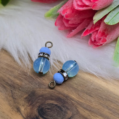 Das Bild zeigt zwei handgefertigte Anhänger mit einer Kombination aus transparenten hellblauen Glasperlen und opaken hellblauen Kristallglasperlen. Ein funkelndes Strassrondell setzt glitzernde Akzente, während antik bronzefarbige Metallelemente dem Design einen charmanten Vintage-Touch verleihen. Die filigranen Details und die harmonische Farbzusammenstellung machen die Anhänger zu einem stilvollen Blickfang für Schmuckkreationen.