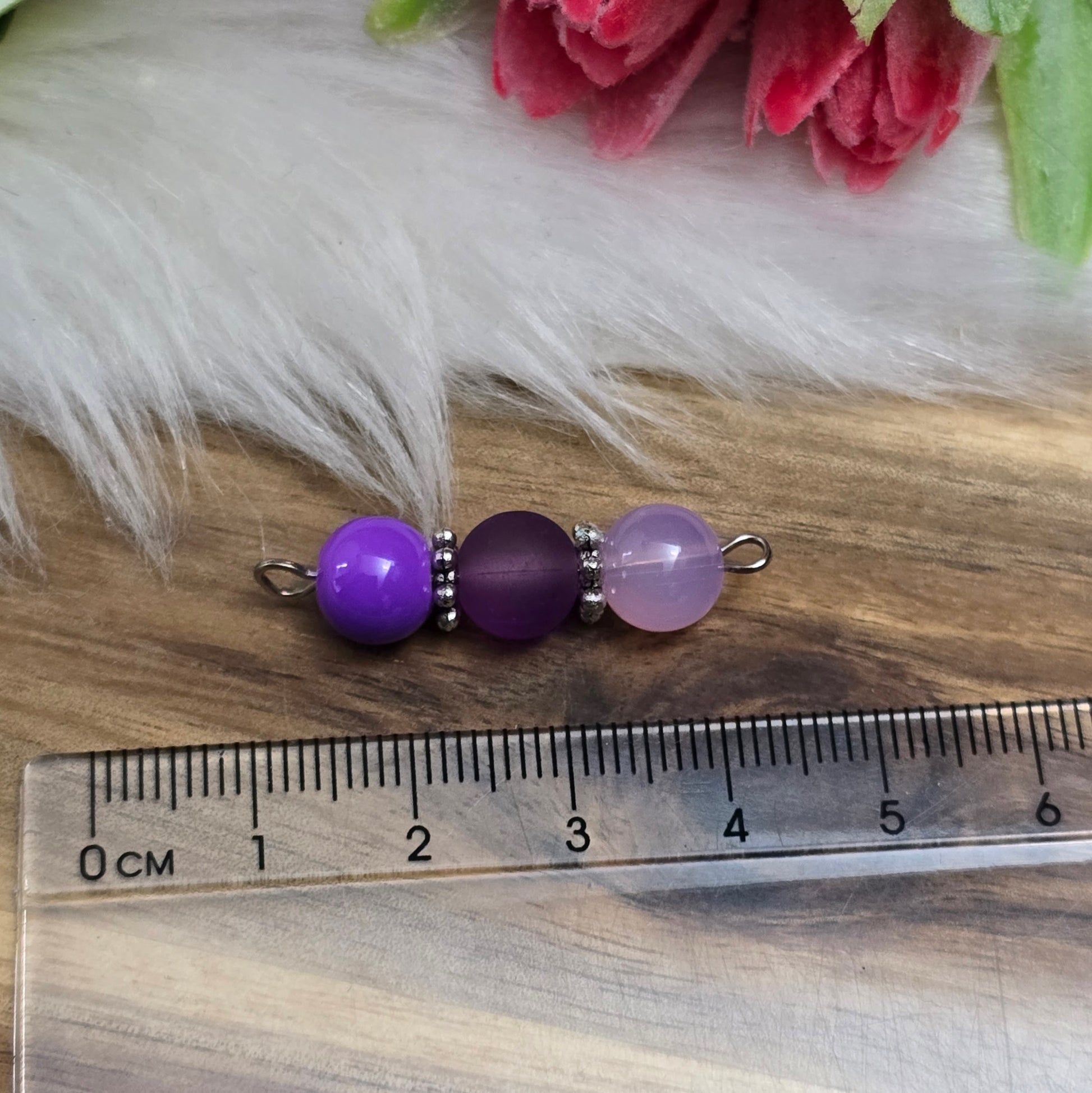 Das Bild zeigt zwei handgefertigte Verbinder mit lila und violetten Glasperlen in unterschiedlichen Ausführungen – eine opake, eine matte und eine milchig transparente. Die Perlen sind harmonisch angeordnet und werden von detailreich verzierten, antik-silberfarbenen Metallelementen eingefasst. Die filigranen Zwischenelemente verleihen den Verbindern eine edle Vintage-Optik. Mit einer Länge von ca. 33mm eignen sie sich perfekt für kreative Schmuckdesigns wie Armbänder, Ketten oder Ohrringe.