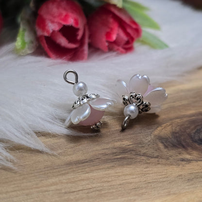Auf dem Bild sind zwei handgefertigte Anhänger in Form von kleinen Blumen zu sehen. Die Anhänger bestehen aus einer eleganten weißen Acrylblüte und einer zarten rosafarbenen Kristallglasperle. Silberfarbige Metallelemente runden das Design ab und verleihen den Anhängern einen romantischen Charme. Mit einer Größe von ca. 18mm sind sie ideal für die individuelle Schmuckgestaltung, sei es für Ketten, Armbänder oder Ohrringe.