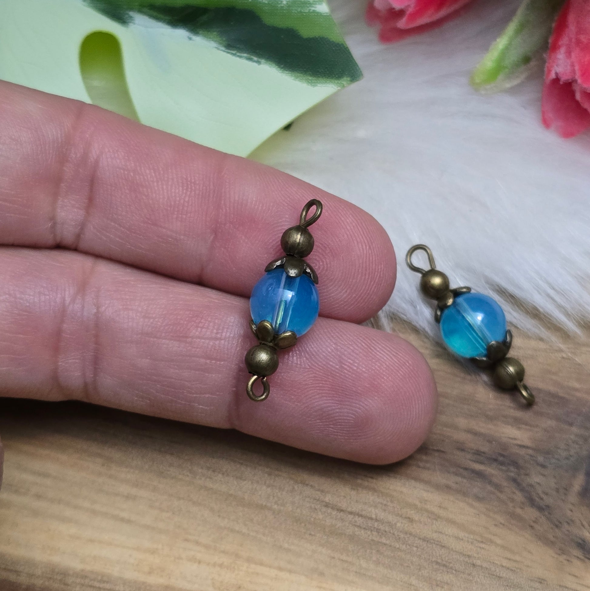 Das Bild zeigt zwei handgefertigte Verbinder mit transparenten, aquablauen Glasperlen, die in antik bronzefarbige Metallelemente eingefasst sind. Die Perlen schimmern sanft und verleihen den Verbindern eine edle Optik. Die filigrane Metallverzierung betont den Vintage-Charakter der Schmuckelemente. Mit einer Länge von ca. 24mm sind sie vielseitig einsetzbar – perfekt für Armbänder, Ketten oder Ohrringe.