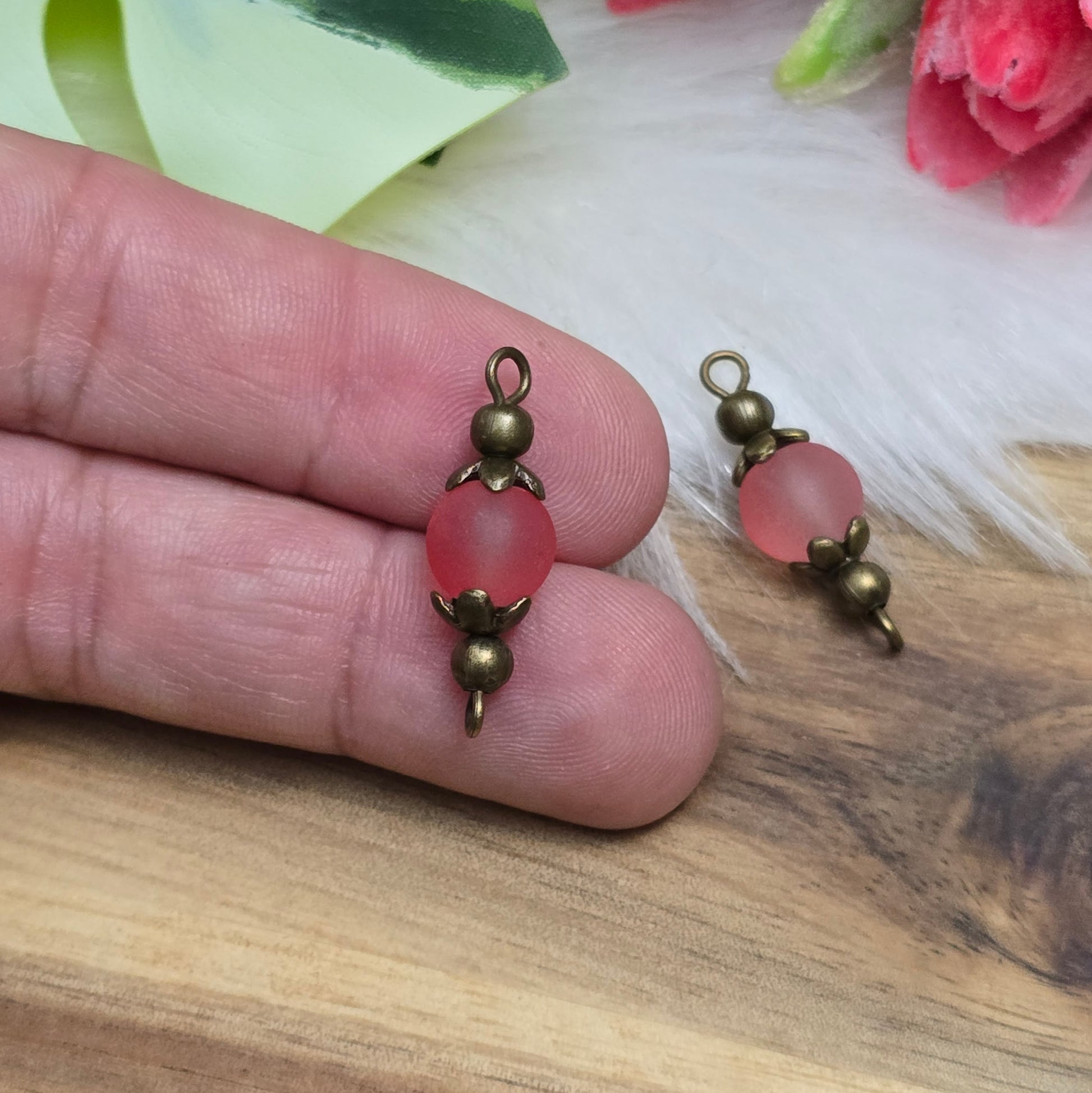 Die Aufnahme zeigt zwei filigrane, handgefertigte Schmuckverbinder mit matten lachsfarbenen Glasperlen. Die sanft schimmernden Perlen sind in kunstvoll verzierten, antik bronzefarbenen Metallelementen eingefasst, die ihnen eine elegante Vintage-Optik verleihen. Die warmen Farbtöne der Glasperlen harmonieren perfekt mit dem dunklen Metall und machen die Verbinder zu einem stilvollen Highlight für Schmuckkreationen.