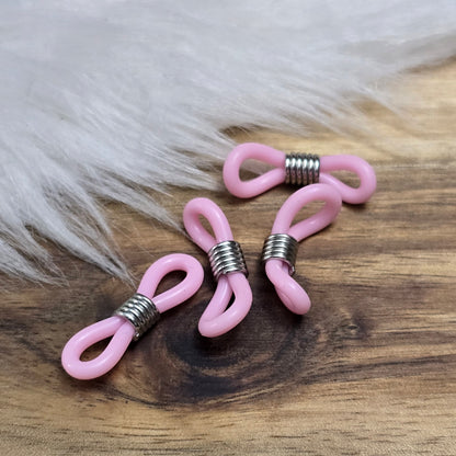 Das Bild zeigt eine Auswahl an Silikonschlaufen für Brillenketten und Maskenhalter in drei Farben: Mint, Rosa und Schwarz. Jede Schlaufe hat eine flexible, weiche Textur und ist so gestaltet, dass sie einfach auf den Bügel der Brille geschoben werden kann. An einem Ende der Schlaufe befindet sich ein kleiner Metallring, der fest angebracht ist, um ein Abrutschen zu verhindern. Die anderen Enden der Schlaufen sind offen, sodass sie mit einem Karabiner oder Biegering leicht an einer Kette befestigt werden kön