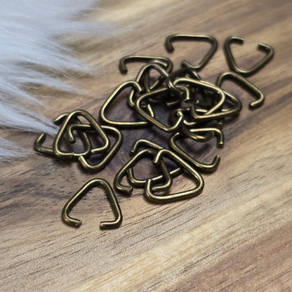 Das Bild zeigt ein Set mit 40 offenen Triangel-Ösen aus antik bronzefarbener Eisenlegierung, die eine Höhe von 9 mm haben. Die Ösen sind in Form eines gleichseitigen Dreiecks gestaltet, wobei eine Seite offen ist, um das Einfügen von Schmuckkomponenten zu erleichtern. Die matte, antik bronzefarbene Oberfläche verleiht den Ösen einen charmanten Vintage-Look, der ideal für verschiedene Schmuckherstellungsprojekte ist. Das Set ist übersichtlich angeordnet und hebt die Vielseitigkeit dieser Ösen hervor, die per