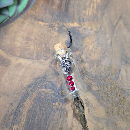 Mini-Glasflasche mit Korken, dekoriert mit Teufelsfratze aus Metall und drei roten Strasssteinen, ideal als Schmuckanhänger oder Sammlerstück.