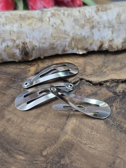 Drei silberne Metall-Haarspangen, davon eine im geöffneten Zustand (Klick-Mechanismus), dekoriert auf einer rustikalen Holzscheibe.