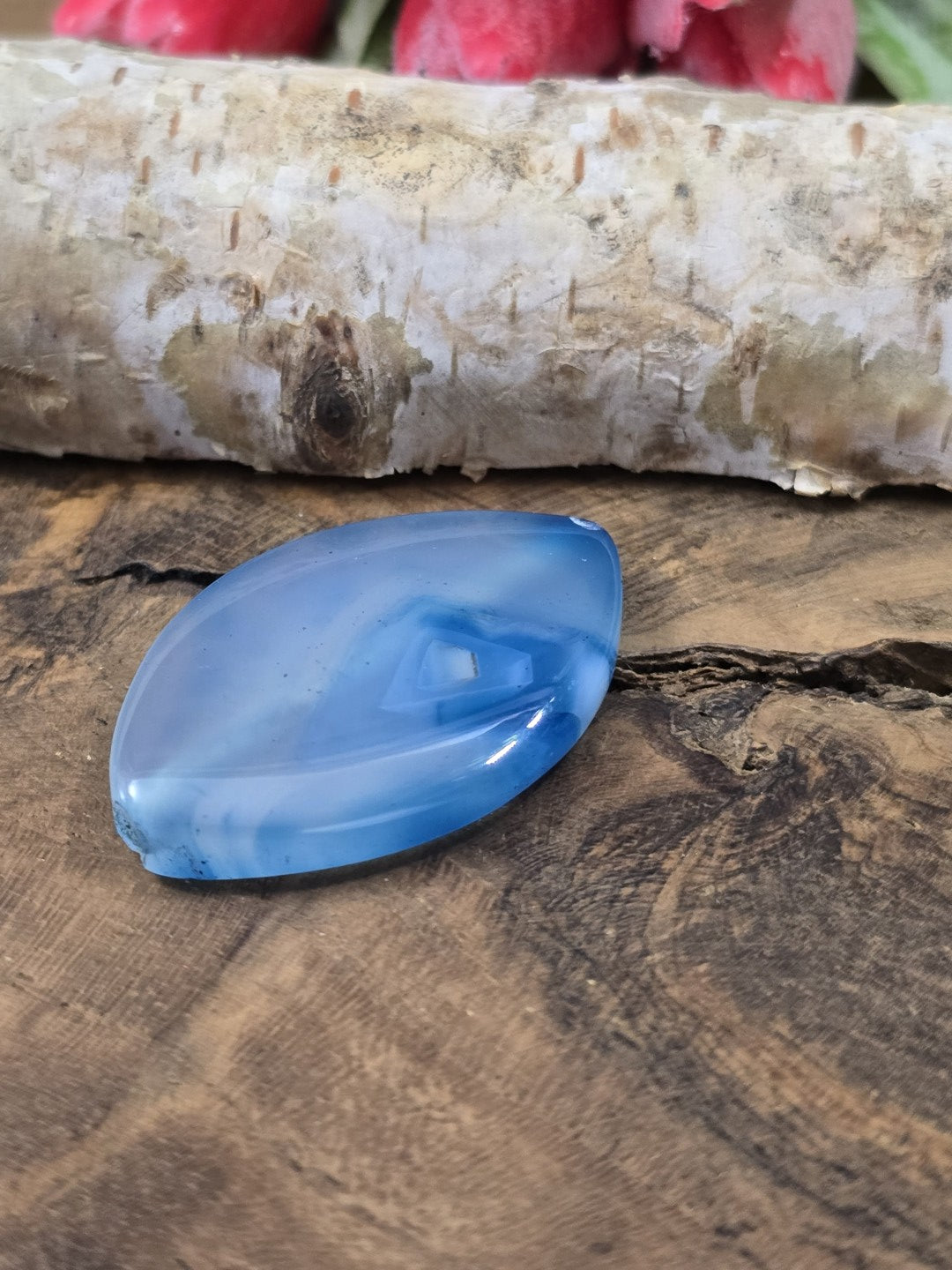 Flach liegende blaue Achat-Perle in Navette-Form auf Holz, um die seitliche Wölbung und den Glanz der Edelsteinperle zu zeigen.